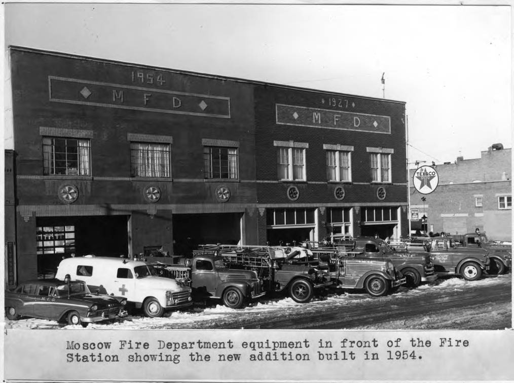 Black-and-white photo of fire trucks and emergency vehicles parked outside a two-story brick fire station. The building has signs reading "1954" and "1927 MFD." A Texaco gas sign is visible in the background.