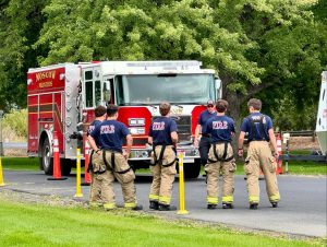 A group of firefighters in uniform and gear stand near a red fire truck labeled "Moscow Volunteers" on a paved road, with green trees and grass in the background.