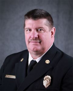 A man in a formal dark uniform with badges and a nameplate poses for a professional portrait against a gray background. He has short dark hair and a mustache.