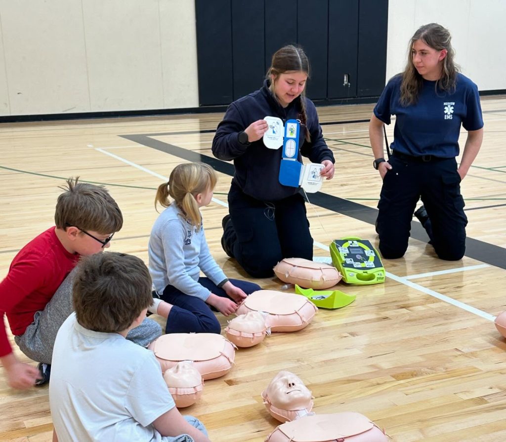 A woman demonstrates how to use an AED device to a group of children seated on a gym floor, with CPR mannequins laid out in front of them. Another instructor looks on attentively.