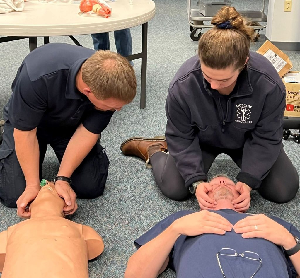 Two people in uniform practice airway management techniques, one on a medical mannequin and the other on a person lying on the floor, in a training room with tables and medical supplies in the background.