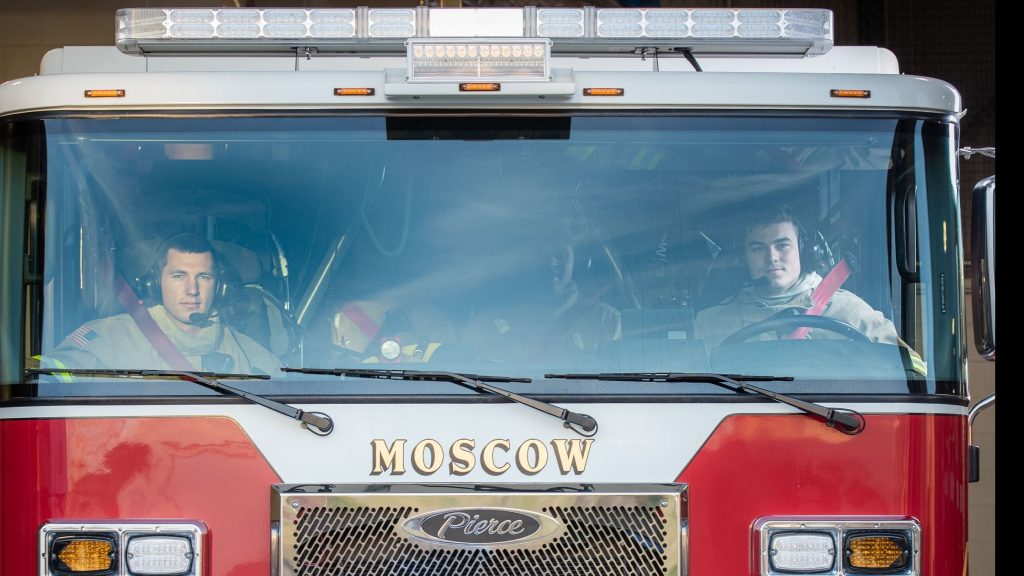 Two firefighters sit inside a red fire truck labeled "Moscow," looking forward through the windshield. Both are wearing headsets and seatbelts, with sunlight reflecting off the glass.