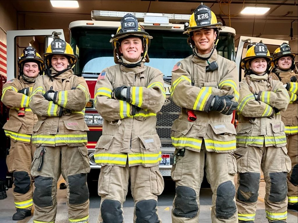 Five firefighters in full gear stand in front of a fire truck with their arms crossed, smiling and posing confidently for the photo in a fire station.