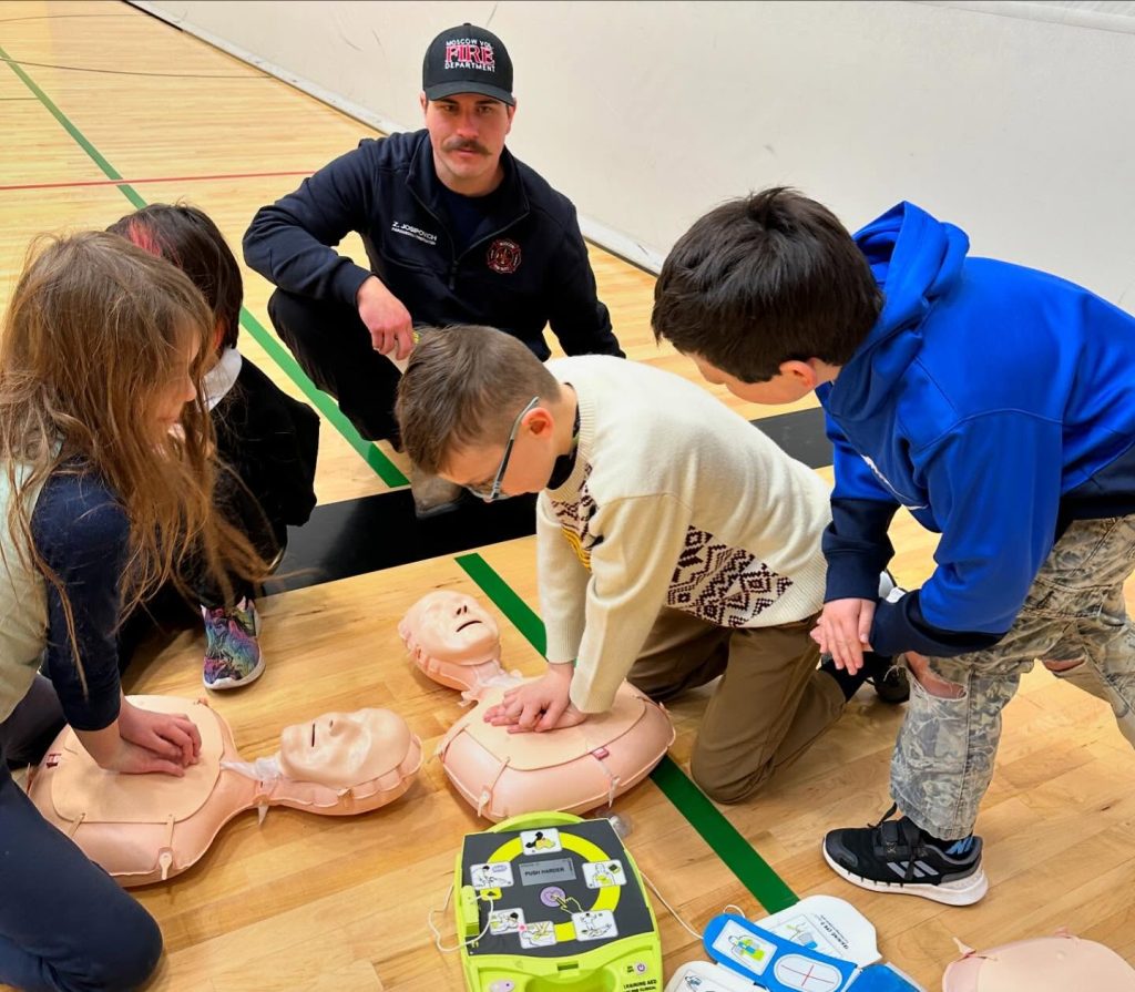 A group of children practice CPR on manikins in a gym while a firefighter supervises. An AED training device is on the floor nearby.