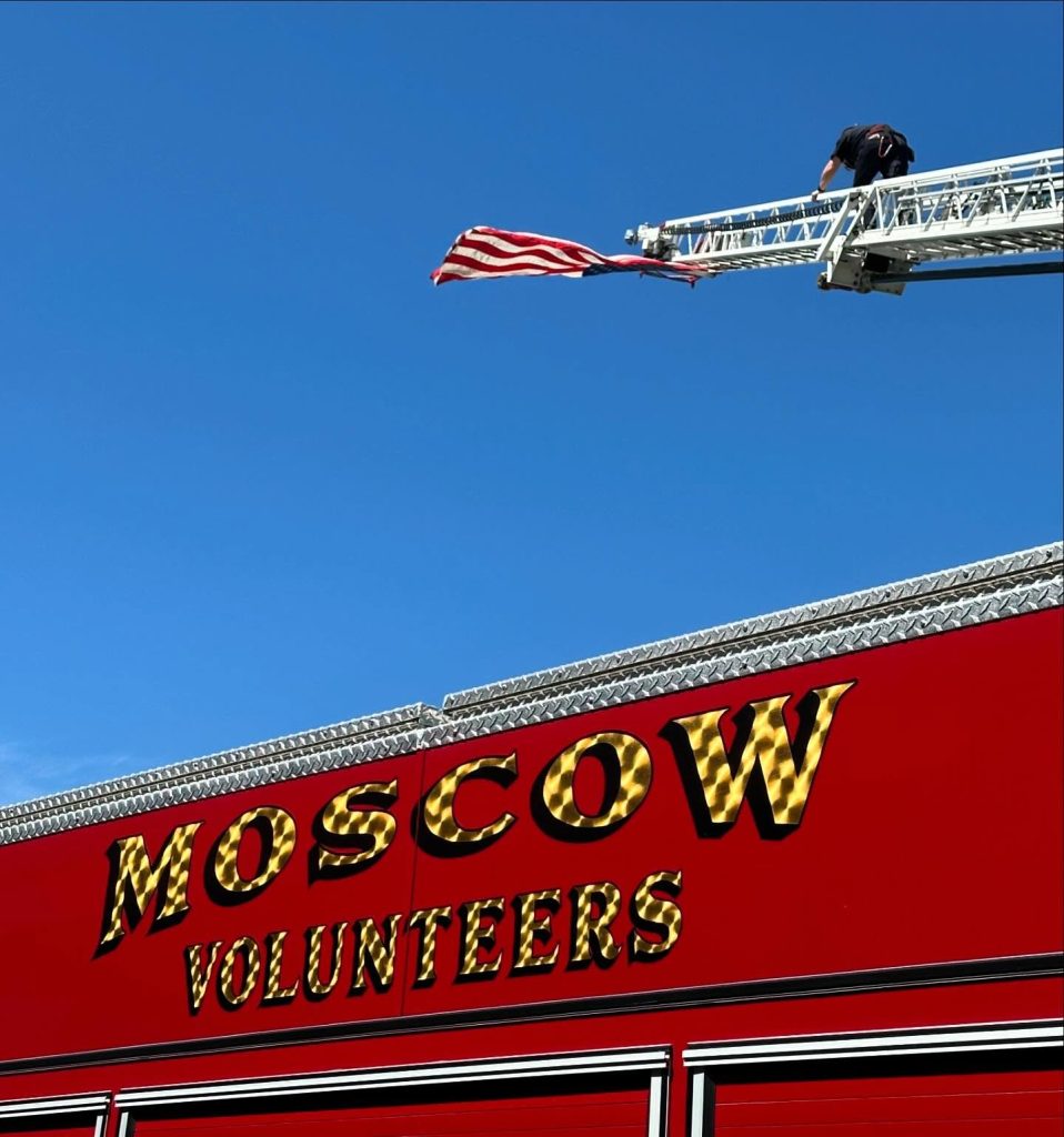 A firefighter raises an American flag atop an extended ladder above a red fire truck labeled "Moscow Volunteers" against a clear blue sky.