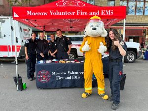 Five firefighters and a person in a fire dog mascot costume stand under a red “Moscow Volunteer Fire and EMS” tent, smiling behind a table with safety materials. One firefighter gives a thumbs-up.