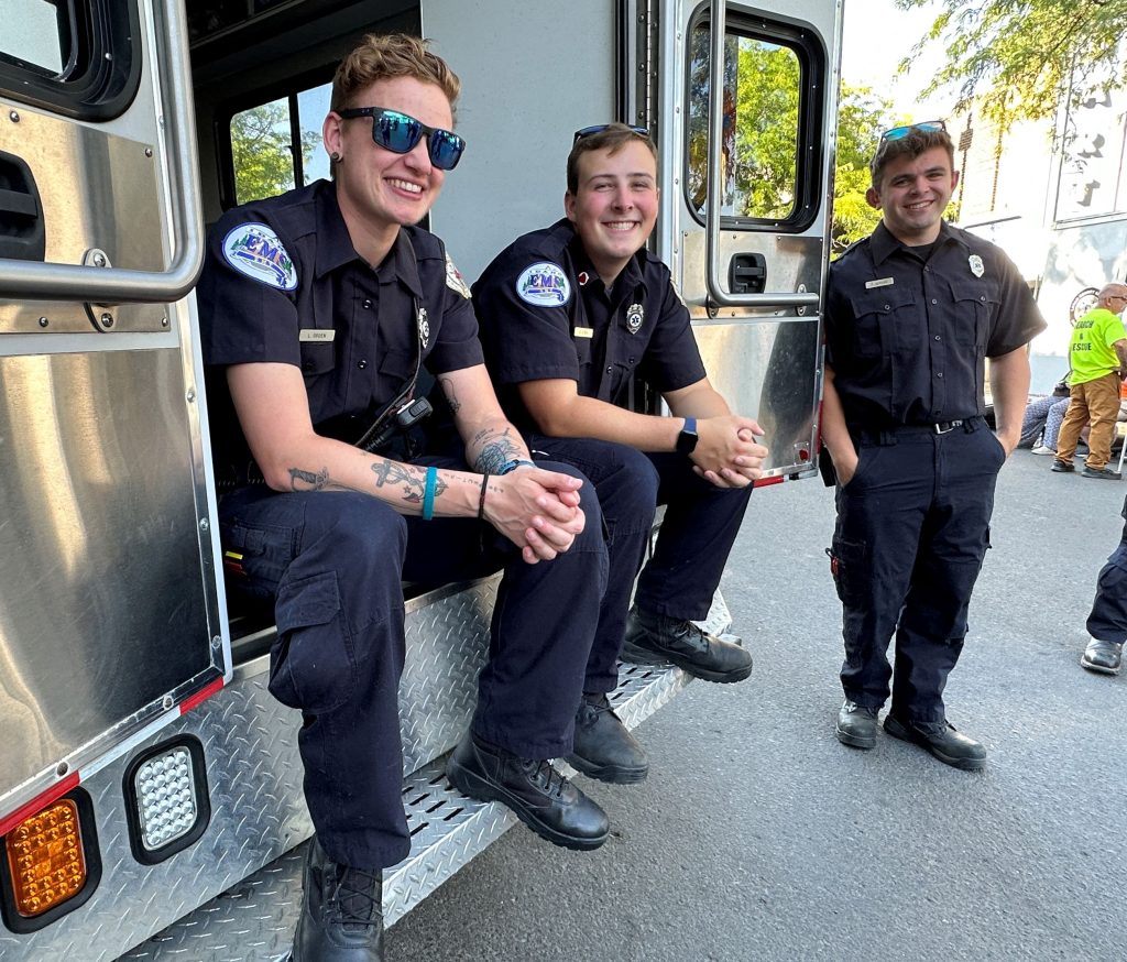 Three smiling emergency medical service workers in uniform sit and stand at the back of an ambulance, enjoying a moment together outdoors on a sunny day.