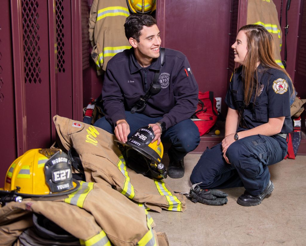 Two firefighters in uniform sit and smile at each other in a locker room, surrounded by firefighting gear, including yellow helmets and tan jackets, with maroon lockers in the background.