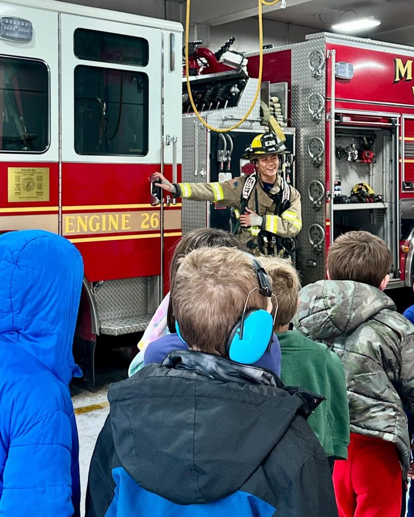 A firefighter in gear stands beside a fire truck, smiling and talking to a group of children wearing colorful jackets, with one child in blue earmuffs, inside a fire station.