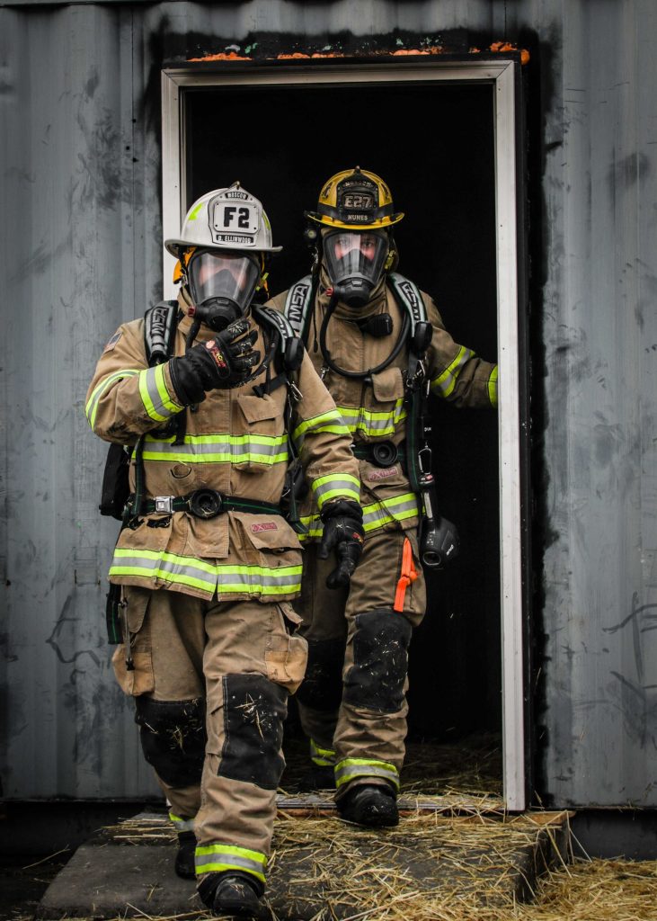 Two firefighters in full gear and breathing masks walk out of a smoky doorway of a gray building. One gestures forward while the other follows, both surrounded by hay and wearing reflective stripes.